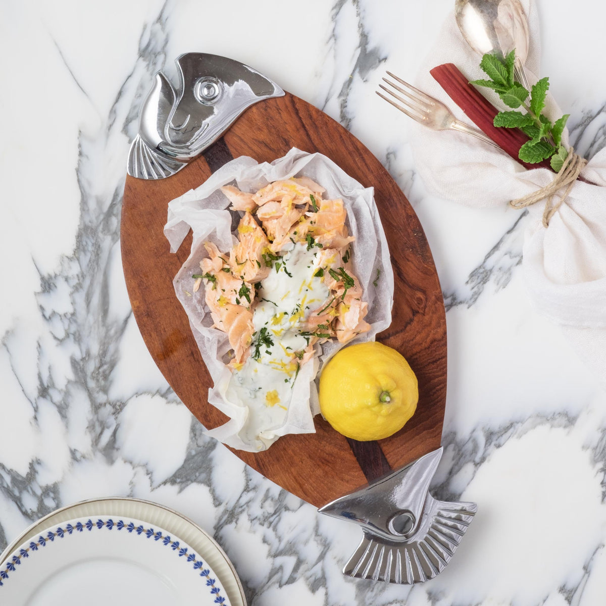 Lachs auf Holzbrett mit angeschnittener Zitrone, daneben Gurke-Rhabarbersalat und ein leeres Wasserglas